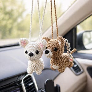 Op de afbeelding: Twee gehaakte hondenauto-bedels die aan de achteruitkijkspiegel van een auto hangen. E&eacute;n is wit met roze oren, de andere is bruin met een kralenstaart die "ROCKY" en een rood hart spelt. De bedels hangen aan witte en bruine koorden.
