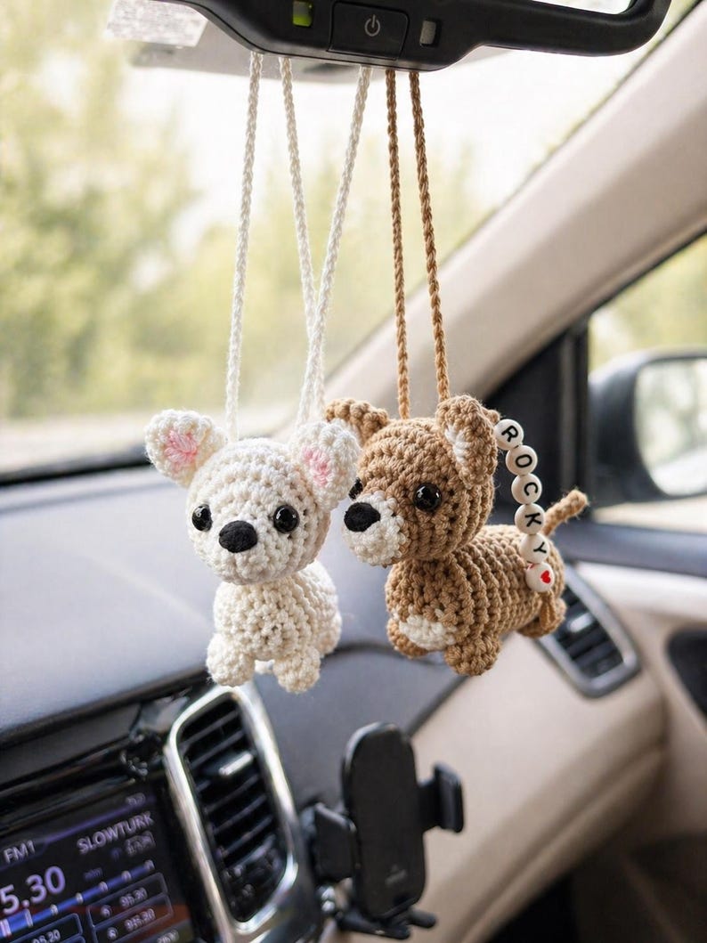 Op de afbeelding: Twee gehaakte hondenauto-bedels die aan de achteruitkijkspiegel van een auto hangen. E&eacute;n is wit met roze oren, de andere is bruin met een kralenstaart die "ROCKY" en een rood hart spelt. De bedels hangen aan witte en bruine koorden.