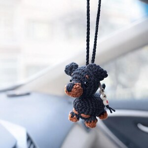 Op de afbeelding: Een handgemaakte zwarte en bruine gehaakte hondenauto-hanger, hangend aan een zwart koord. De hond heeft bruine accenten op zijn poten en snuit. Een kleine bedel met witte kralen en een rood hart is bevestigd.