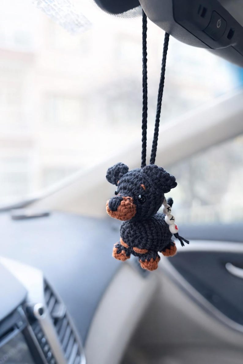 Op de afbeelding: Een handgemaakte zwarte en bruine gehaakte hondenauto-hanger, hangend aan een zwart koord. De hond heeft bruine accenten op zijn poten en snuit. Een kleine bedel met witte kralen en een rood hart is bevestigd.
