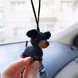 Op de afbeelding: Een gehaakt zwart en bruin teckel hondenornament. De hond heeft grote floppy oren, een lang lichaam en korte poten. Het is opgehangen aan een zwart koord, geschikt om in een auto te hangen of als decoratief item.