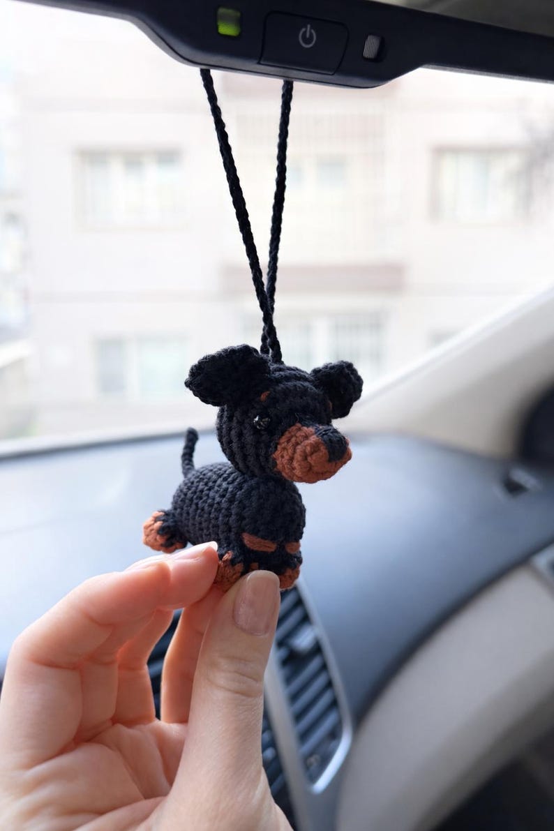 Op de afbeelding: Een gehaakt zwart en bruin teckel hondenornament. De hond heeft grote floppy oren, een lang lichaam en korte poten. Het is opgehangen aan een zwart koord, geschikt om in een auto te hangen of als decoratief item.