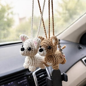 Op de afbeelding: Twee gehaakte hondenauto-bedels hangen aan de achteruitkijkspiegel van een auto. E&eacute;n is wit met roze oren, de andere is bruin. De bruine hondenbedel heeft een kleine houten kraal met "ROCKY". Het dashboard en een telefoonhouder zijn zichtbaar.