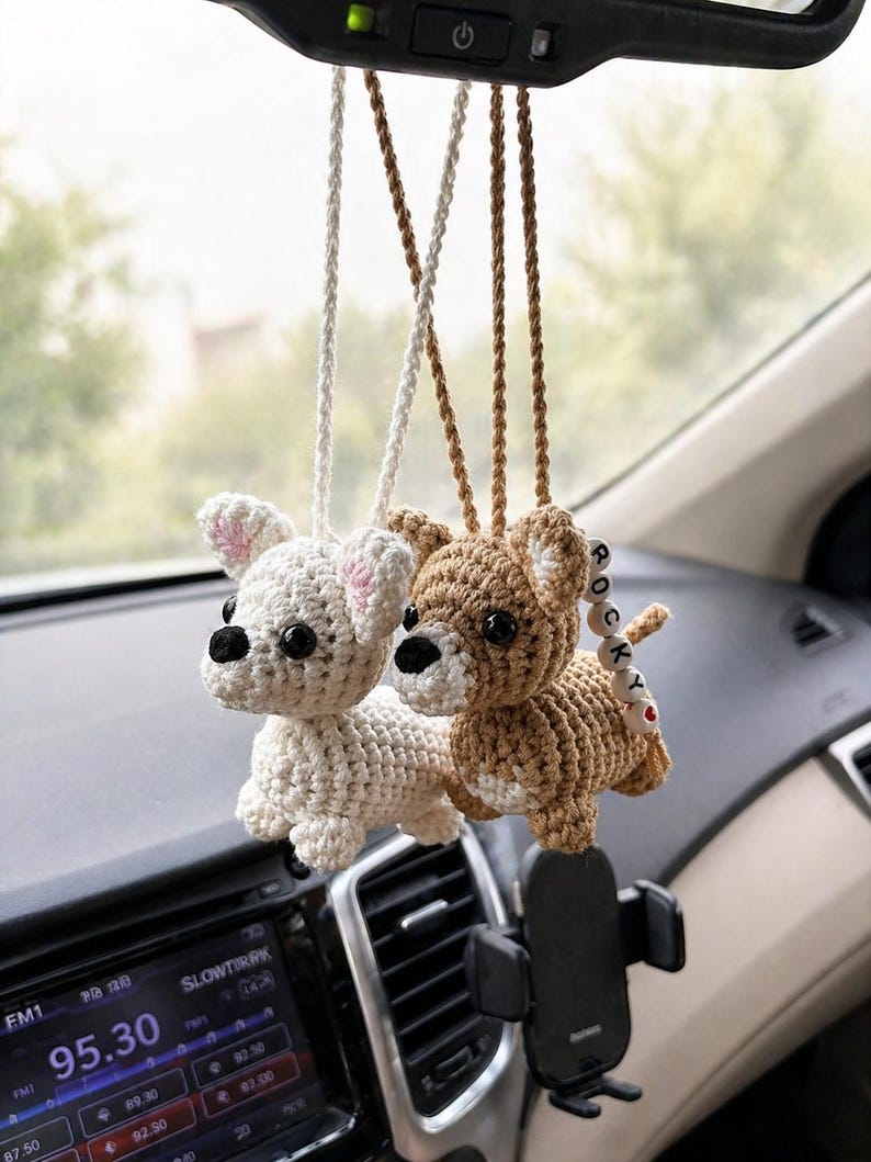 Op de afbeelding: Twee gehaakte hondenauto-bedels hangen aan de achteruitkijkspiegel van een auto. E&eacute;n is wit met roze oren, de andere is bruin. De bruine hondenbedel heeft een kleine houten kraal met "ROCKY". Het dashboard en een telefoonhouder zijn zichtbaar.