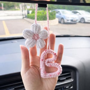 May include: A handmade crochet car accessory featuring a white five-petaled flower with a pink center and a light pink letter B with small pink accents, both suspended from a matching pink yarn string designed for hanging from a rearview mirror.