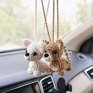 Op de afbeelding: Twee gehaakte hondenauto-hangers die aan de achteruitkijkspiegel van een auto hangen. E&eacute;n is wit met roze accenten, de andere is bruin. Beide hebben zwarte ogen en zijn bevestigd met bijpassende koorden.
