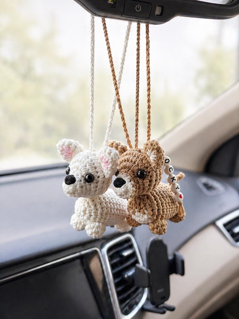 Op de afbeelding: Twee gehaakte hondenauto-hangers die aan de achteruitkijkspiegel van een auto hangen. E&eacute;n is wit met roze accenten, de andere is bruin. Beide hebben zwarte ogen en zijn bevestigd met bijpassende koorden.