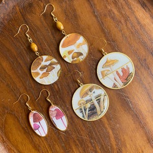 May include: A collection of five pairs of earrings with mushroom designs. The earrings feature gold-colored hooks and circular or oval pendants with various mushroom illustrations in shades of brown, red, and white. Some earrings have wooden bead accents.