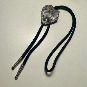 May include: A bolo tie featuring a silver-toned metal wolf head centerpiece attached to a black braided cord. The cord ends are finished with matching silver-toned metal tips that have a ribbed texture and a rounded, smooth bottom edge.