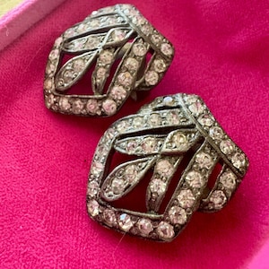 May include: A pair of silver-toned metal clip-on earrings featuring a leaf-inspired openwork design. Each earring is encrusted with numerous small, clear, round-cut rhinestones that cover the entire surface of the metal frame in a dense pattern.