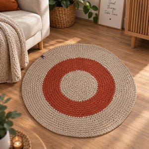 Könnte beinhalten: Ein runder, handgehäkelter Teppich mit konzentrischem Design, bestehend aus einer beigen Mitte, einem breiten rostorangen Mittelring und einem beigen Außenrand. Der Teppich hat eine dicke, strukturierte Garnbeschaffenheit und ein kleines schwarzes Etikett.