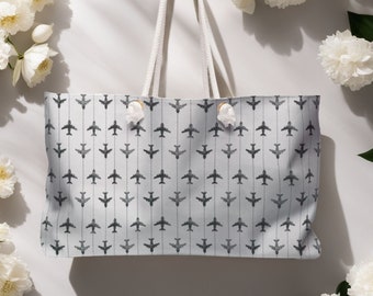 May include: A white tote bag with a gray airplane pattern. The bag has white rope handles and is surrounded by white flowers.