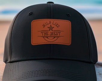 Pode incluir: Boné de beisebol preto com um remendo de couro marrom que diz "Wild Like The West" com uma estrela e uma ferradura.