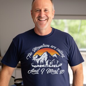 Może przedstawiać: Koszulka w kolorze granatowym z grafiką gór i zachodu słońca. Tekst na koszulce brzmi "The Mountains are calling And I Must go".