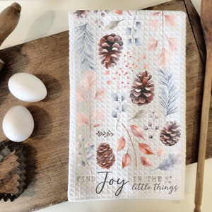 May include: White waffle-weave kitchen towel with a watercolor design of pinecones, leaves, and berries in shades of brown, pink, and blue. The towel has the text "Find Joy in the little things" in black script.