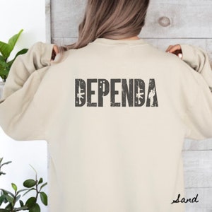 May include: A beige sweatshirt with the word "DEPENDA" printed on the back in black with a distressed look. The word is surrounded by small images of guns and soldiers.