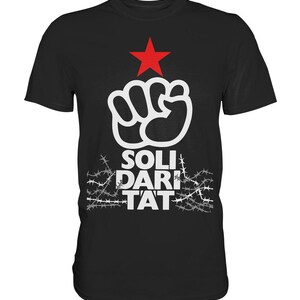 Pode incluir: Camiseta preta com um punho branco e uma estrela vermelha acima. O texto "SOLIDARITAT" está abaixo do punho, rodeado de arame farpado.