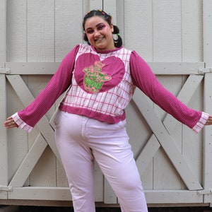 May include: A person wearing a pink and white plaid long-sleeved shirt with a green and pink heart-shaped patch that says "PRESENCE". The person is wearing light pink pants.
