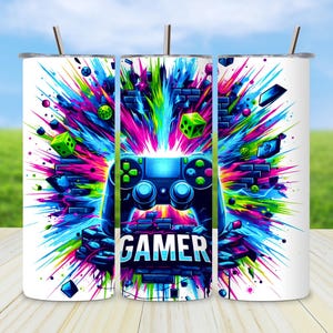 May include: White tumbler with a vibrant, colorful design featuring a blue video game controller and the word "GAMER" in white. The design includes splashes of pink, green, blue, and purple, with a straw.