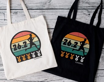 May include: Two reusable tote bags with a retro design featuring a runner and the text "26.2" with medals. The bags are 15 inches by 16 inches and are made of cotton canvas. One bag is white and the other is black.
