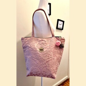 Może przedstawiać: Różowa torba typu tote z kwiatowym wzorem i zamkiem błyskawicznym. Do suwaka przymocowany jest mały różowo-brązowy pompon.