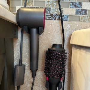 May include: A black hair dryer and a black and pink Revlon hair styling brush are mounted on a wall. The hair dryer has a pink interior and is plugged into an outlet. The styling brush has pink bristles.