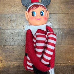 May include: A vintage Christmas elf doll with a Mickey Mouse ear hat. The doll has a red and white striped outfit, black shoes, and a white collar. The elf has rosy cheeks, blue eyes, and red hair.