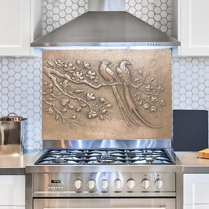 May include: A bronze-coloured metal backsplash with an embossed design of two birds perched on a branch with flowers. The backsplash is installed above a stainless steel stove with a black hob.
