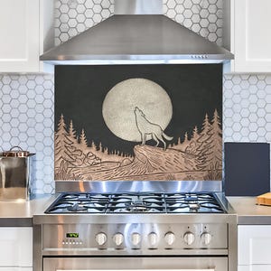 Puede incluir: Una estufa de acero inoxidable con un protector contra salpicaduras negro y cobre que presenta un lobo aullando a la luna en un entorno de bosque.