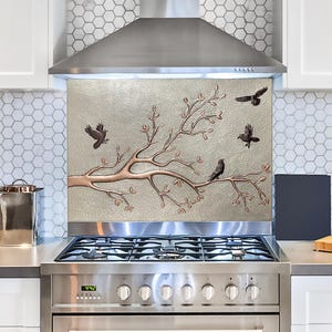 May include: A silver and copper kitchen backsplash with a tree branch design and birds. The backsplash is installed above a stainless steel stove with a black cooktop.