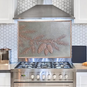 May include: A decorative kitchen backsplash featuring a detailed relief of pine branches and pinecones in shades of brown and copper against a muted green background. The backsplash is framed in a copper-colored border and is mounted above a stainless steel gas range.