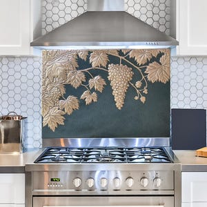 May include: A kitchen backsplash with a gold-colored grape vine design against a teal background. The backsplash is installed above a stainless steel gas range and below a stainless steel range hood. White cabinets and a white and gray hexagon tile pattern are visible.