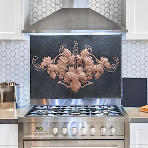 May include: A stainless steel range hood with a copper-colored grapevine design on the backsplash. The range hood has a black glass cooktop with four burners and a stainless steel oven below.