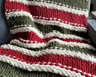 Könnte beinhalten: Eine gestreifte Strickdecke in Rot, Weiß und Grün. Die Decke ist aus dickem Garn gefertigt und hat eine weiche, kuschelige Textur.