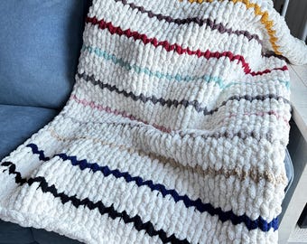 May include: A white chunky knit throw blanket with colourful stripes in shades of red, blue, yellow, and brown. The blanket is draped over a blue sofa.