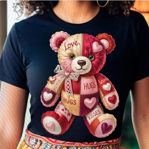May include: A navy blue t-shirt featuring a colorful teddy bear design. The bear is a patchwork of pink, red, and cream-colored fabrics, with hearts and the words "Love," "Hugs," and "Kisses" on it. A bow is at the bear's neck.
