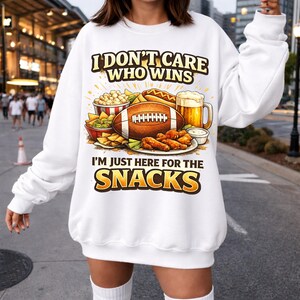 May include: White sweatshirt with a colorful graphic of football-themed snacks, including a football, hot dog, beer, and chicken wings. Text on the shirt reads "I DON'T CARE WHO WINS I'M JUST HERE FOR THE SNACKS."