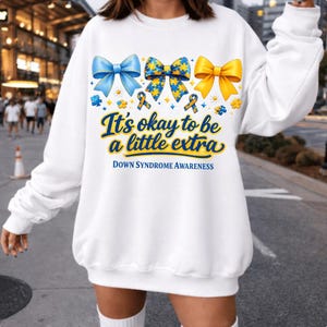 Può includere: Felpa bianca con tre fiocchi colorati: blu, giallo e un motivo blu e giallo. Il testo "It's okay to be a little extra" e "Down Syndrome Awareness" è stampato sotto. Il design include pezzi di puzzle e nastri.