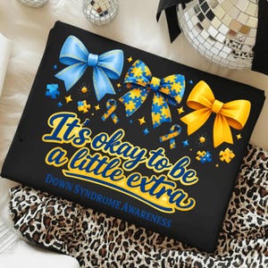 May include: Black t-shirt with the text "It's okay to be a little extra" and "Down Syndrome Awareness" in yellow and blue. The shirt features blue and yellow bows, puzzle pieces, and ribbons.