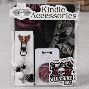 May include: Kindle accessories set in a clear package. Includes a keychain with black, white, and clear beads, a rose-themed pop socket, and stickers with a skull and rose design. The package has the text "Kindle Accessories" and "In my dark Romance Era".
