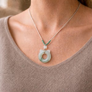 May include: A jade cat pendant necklace. The pendant is a light green color with a circular design and cat ear details. The necklace has a silver chain and a green and white accent. The necklace is worn on a person.