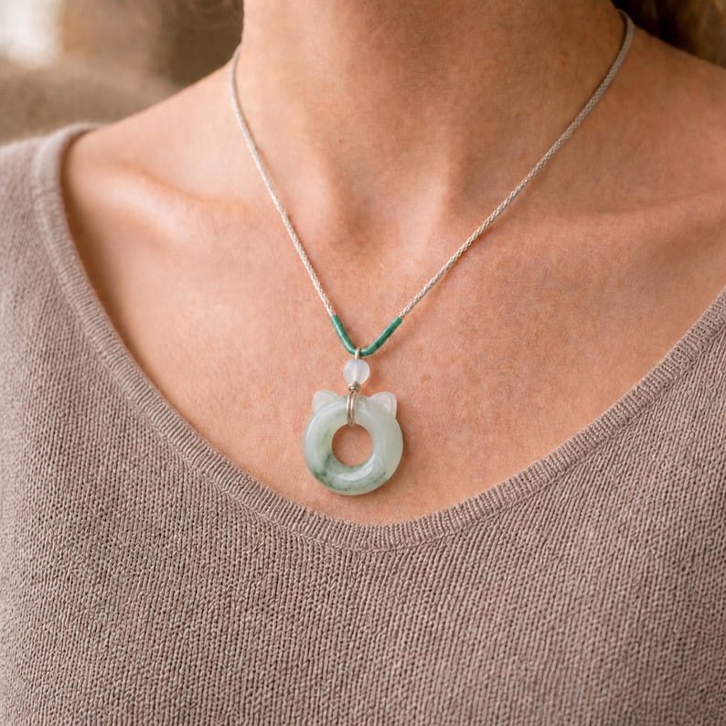 May include: A jade cat pendant necklace. The pendant is a light green color with a circular design and cat ear details. The necklace has a silver chain and a green and white accent. The necklace is worn on a person.