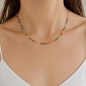 May include: A delicate necklace featuring a single strand of small, multi-colored faceted beads in shades of red, orange, yellow, green, blue, and clear. The beads are spaced evenly along a thin, metallic chain that rests against the collarbone.