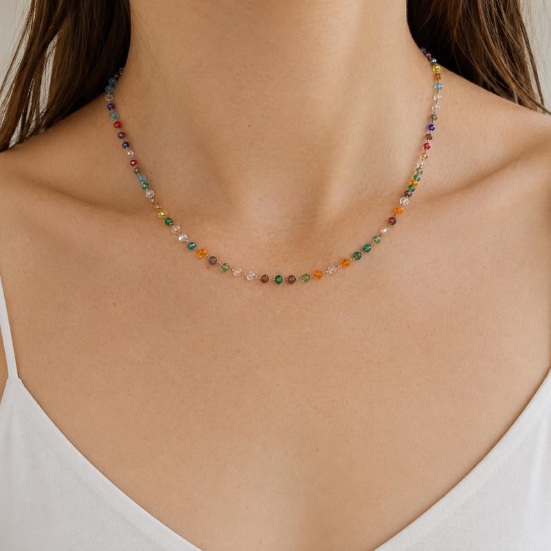 May include: A delicate necklace featuring a single strand of small, multi-colored faceted beads in shades of red, orange, yellow, green, blue, and clear. The beads are spaced evenly along a thin, metallic chain that rests against the collarbone.