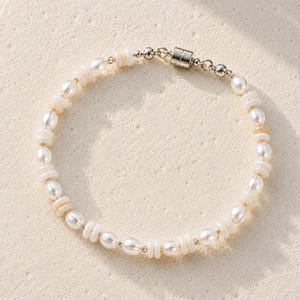 May include: A beaded bracelet featuring alternating white oval pearls and small, flat, circular off-white shell beads. The bracelet is finished with a polished silver-toned metal cylindrical magnetic clasp and small round silver-toned metal spacer beads.