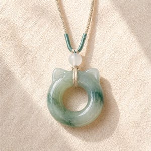 May include: A jade pendant necklace featuring a cat-shaped design with a circular center. The pendant is a mix of green and white hues, suspended from a beige cord with a green accent. A white bead is positioned above the pendant.