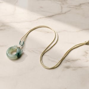 May include: A jade pendant necklace featuring a circular jade stone with a cat-like design. The stone is a mix of green and white, and is strung on a beige cord. The necklace is displayed on a marble surface.