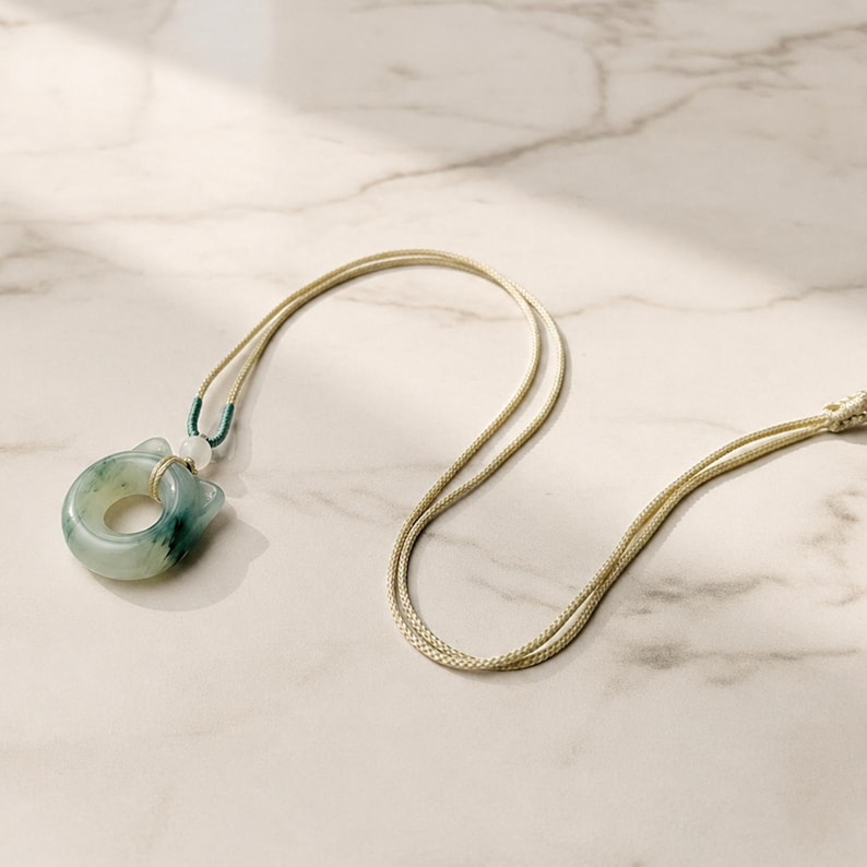 May include: A jade pendant necklace featuring a circular jade stone with a cat-like design. The stone is a mix of green and white, and is strung on a beige cord. The necklace is displayed on a marble surface.
