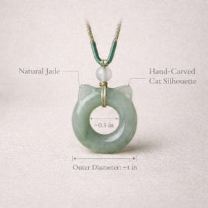May include: A natural jade pendant with a hand-carved cat silhouette. The pendant is approximately 1 inch in outer diameter and has a 0.5-inch inner diameter. It hangs from a green and beige cord.