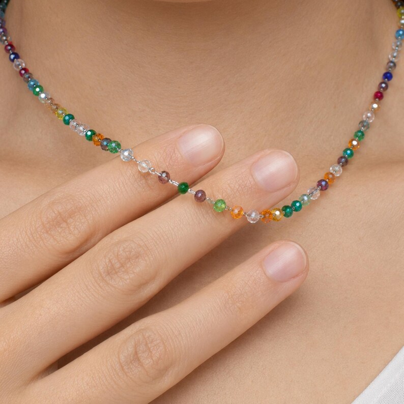 May include: A delicate necklace featuring a single strand of small, multi-colored faceted beads in shades of red, orange, yellow, green, blue, and clear. The beads are strung on a thin, metallic chain that rests against the neck, creating a vibrant look.